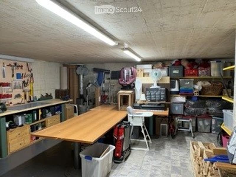 A well-organized workshop or garage space with a large wooden worktable, various tools on the walls, storage bins, a computer setup, and a small refrigerator.