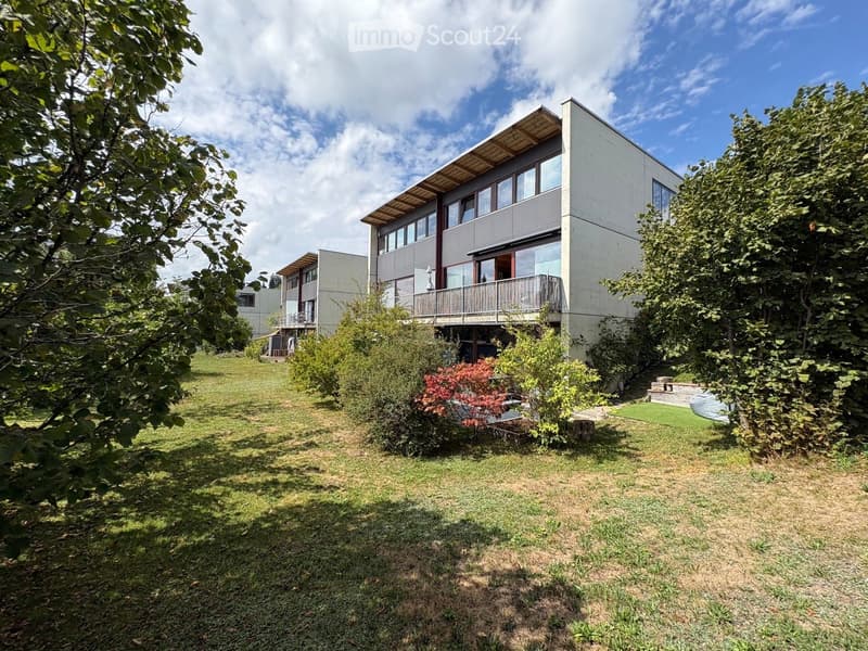 Modern two-story house with large windows and a balcony, surrounded by a well-maintained lawn and trees.