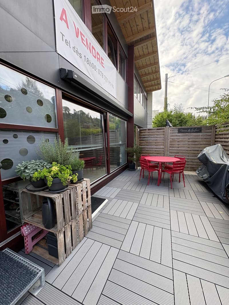 Wooden deck with a red table and chairs, potted plants, and a covered grill. The deck is adjacent to a building with a sign reading "A VENDRE" and a partially visible interior.