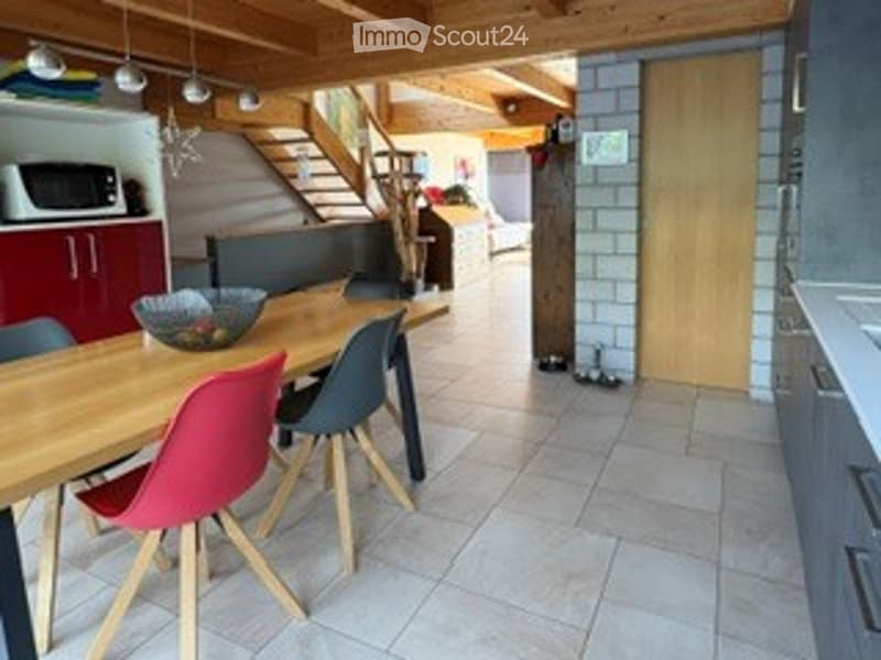 Bright, modern dining area with a wooden dining table, colorful chairs, and a microwave in the background. The space features exposed wooden beams and a staircase leading to another level.