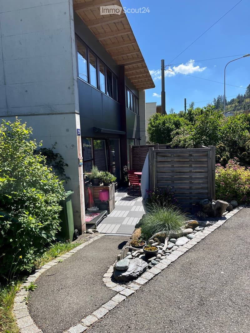 Outdoor area with a wooden deck, potted plants, a small table and chairs, a wooden fence, and a pathway lined with rocks and plants.