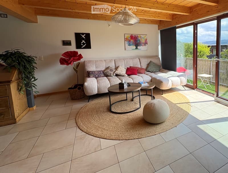 Modern living room with a large sectional sofa, coffee table, ottoman, and floor lamp. The room features a view of the outdoors through large windows and a sliding glass door.