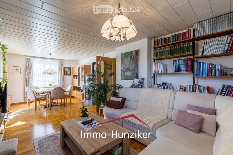 Bright living room with wooden flooring, large windows, a white sofa with throw pillows, a wooden coffee table, dining area with chairs, bookshelf filled with books, and decorative plants.