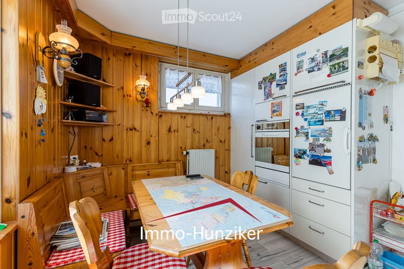 Cozy kitchen with wooden walls and ceiling, featuring a dining table with chairs, a refrigerator covered in photos, and a lamp hanging from the ceiling.