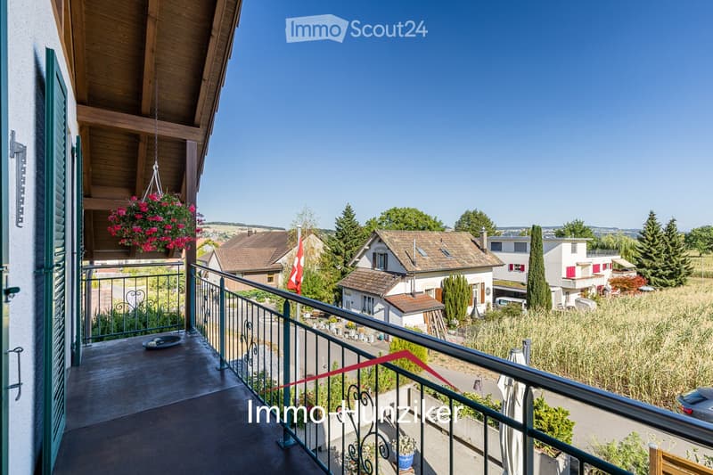 Balcony with wooden floor, metal railing, and hanging flower basket. Offers a view of a residential neighborhood with houses, trees, and a clear blue sky.