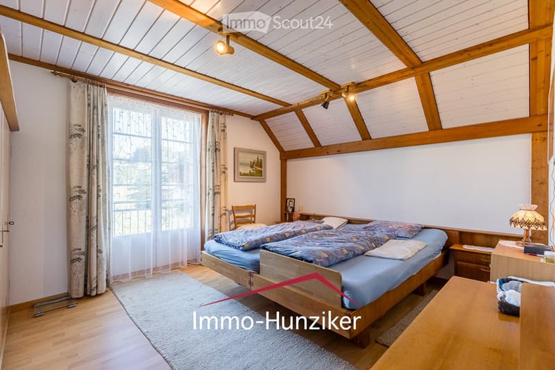 Cozy attic bedroom with wooden beams, large window with curtains, double bed with blue bedding, nightstand, chair, and a small table with a lamp.