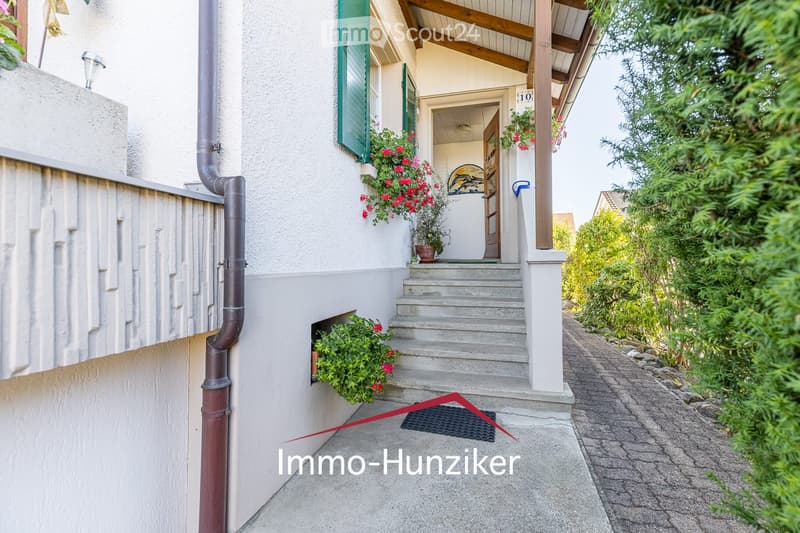 A two-story house with a raised ground floor, featuring a covered entrance with wooden beams, green shutters, and flower boxes on the windows. The pathway is paved, and there is a small garden area with greenery.