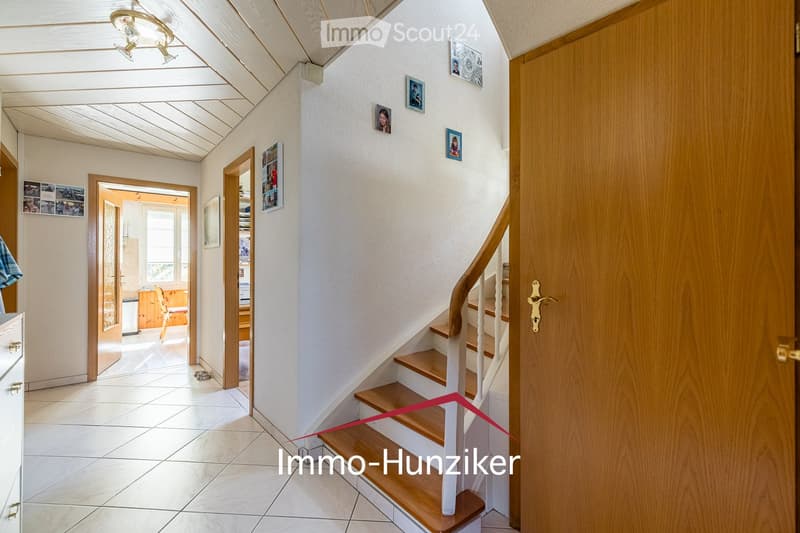 Bright hallway with wooden staircase, white walls, and several doors leading to other rooms. The ceiling has exposed wooden beams, and the floor is tiled.