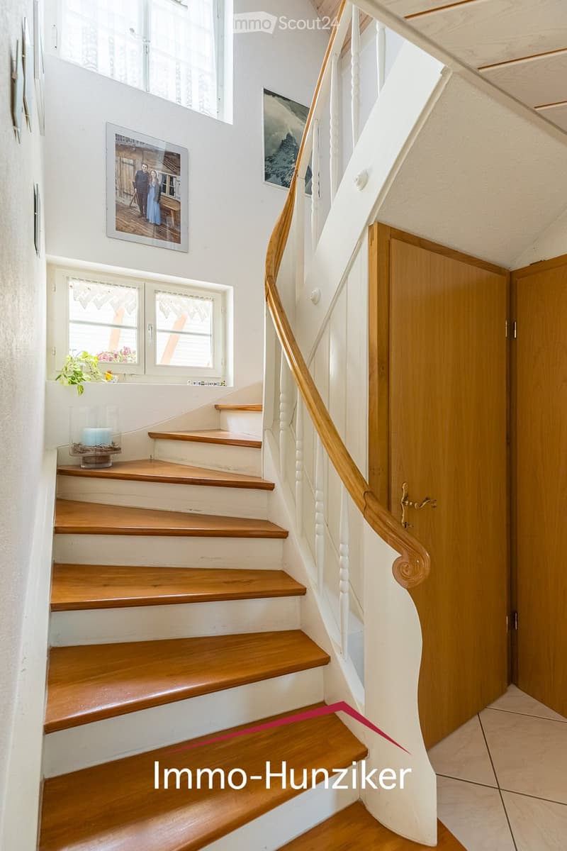 Spiral staircase with wooden steps and white balustrade, leading to a landing area with white walls, framed pictures, and a small window with a plant on the windowsill.
