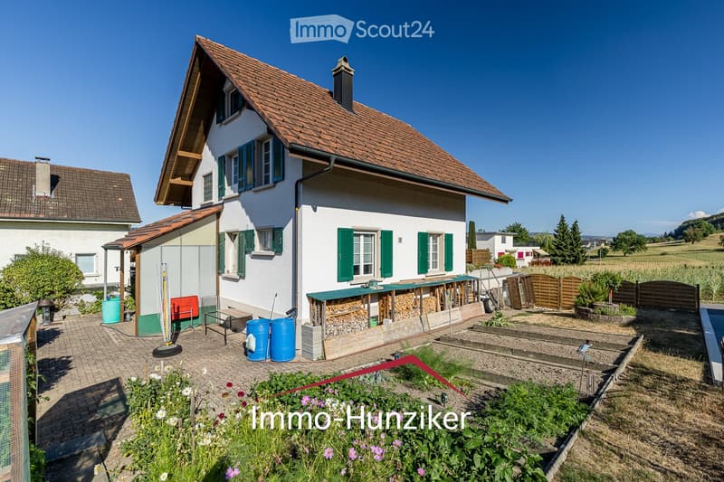 Two-story house with white walls, green shutters, and a red-tiled roof. The house features a garden area with raised beds, a wooden fence, and a small outdoor seating area with a table and chairs.