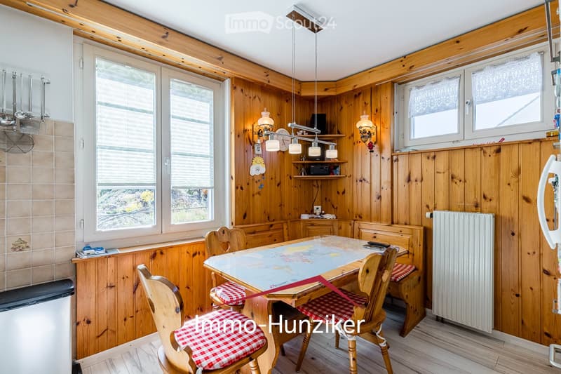 Cozy kitchen with wooden walls and ceiling, featuring a dining table with four chairs, a radiator, and a shelf with various items.