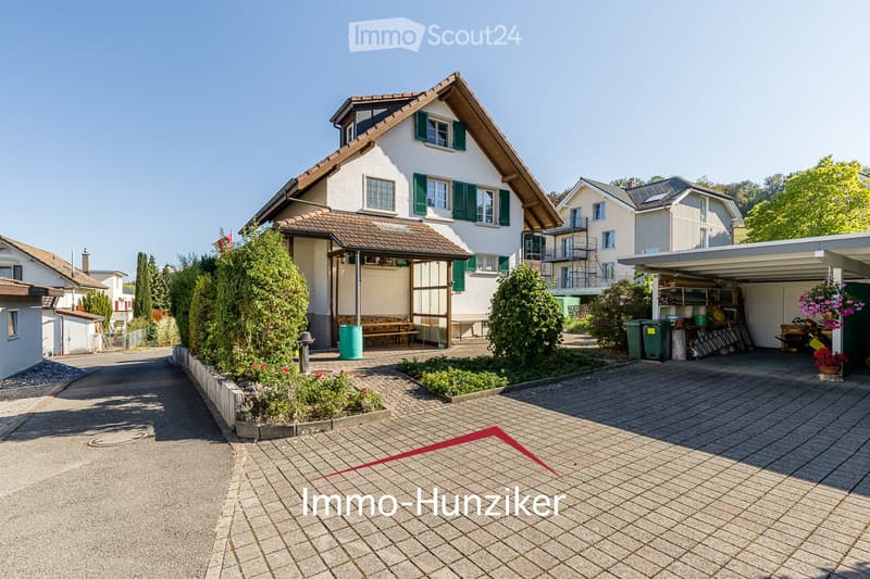 Two-story house with white walls, green shutters, and a tiled roof. The house features a covered porch area with a bench and a small garden bed. A paved driveway leads to a garage with storage items and a hanging basket with flowers.