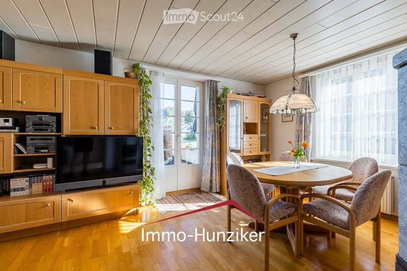 Bright living room with wooden flooring, large windows, a round dining table with chairs, a TV cabinet with a television, and a hanging lamp.