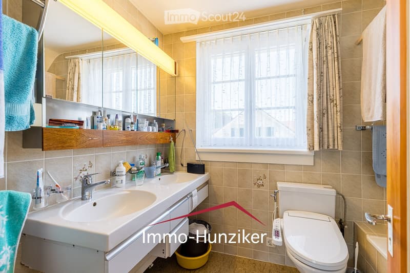 Bathroom with beige tiles, a double sink, a toilet, a mirror, and a window with a curtain. The sink has a faucet, and there are various toiletries and a towel visible.
