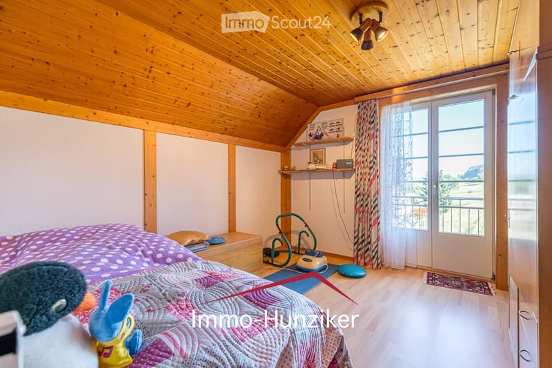 Cozy attic bedroom with wooden beams, a bed with a colorful quilt, a small shelf with books and a mirror, exercise bike, and a large window with a view.