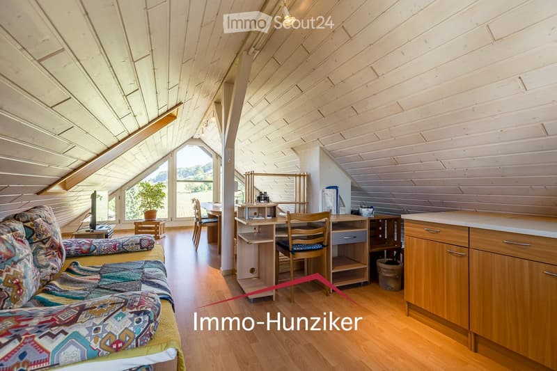 Bright, wooden-floored room with a slanted wooden ceiling, featuring a sofa, small table, chairs, kitchen area with cabinets and counter, and a potted plant near the window.