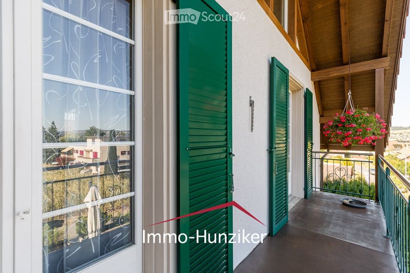 Balcony with green shutters, wooden ceiling, metal railing, and a hanging flower basket with pink flowers.