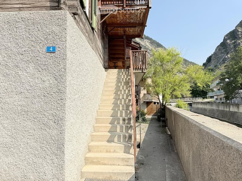Narrow stone-paved pathway leading to a wooden staircase and balcony, with a stone wall on one side and a wooden building on the other.