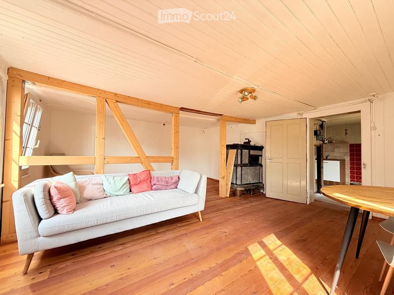 A cozy living room with wooden floors, exposed wooden beams, a grey sofa with colorful cushions, a wooden coffee table, and a fireplace. The room has large windows and a door leading to another area.