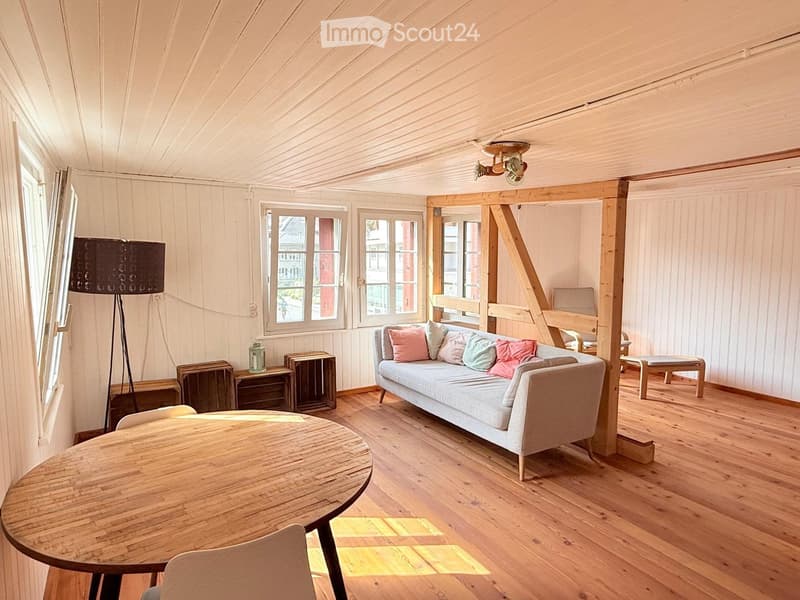 Bright living room with wooden floors, white paneled walls, and large windows. Features a light grey sofa with colorful cushions, a round wooden table, and a tall floor lamp.