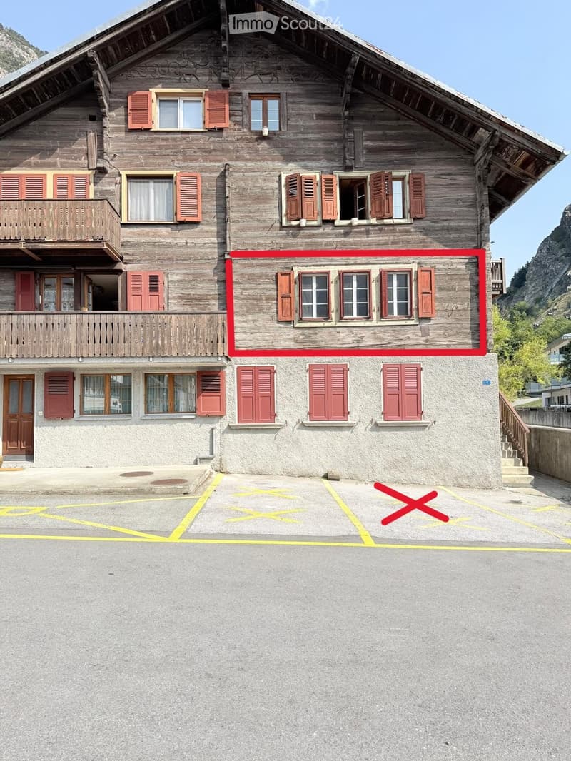 Traditional wooden house with multiple stories, wooden balconies, and red shutters. The ground floor has a stone facade and a parking area.