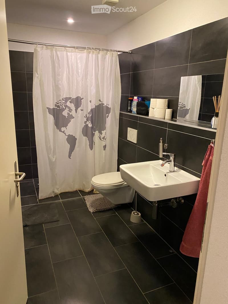 Modern bathroom with dark tiled walls and floor, white toilet, sink, and mirror. Features a world map shower curtain and a red towel hanging on the side.