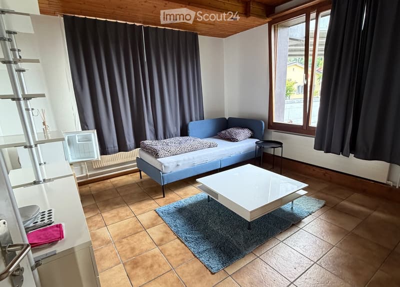 A furnished room with a sofa, coffee table, small stool, refrigerator, and a curtain covering a window. The room has tiled flooring and a blue rug under the coffee table.