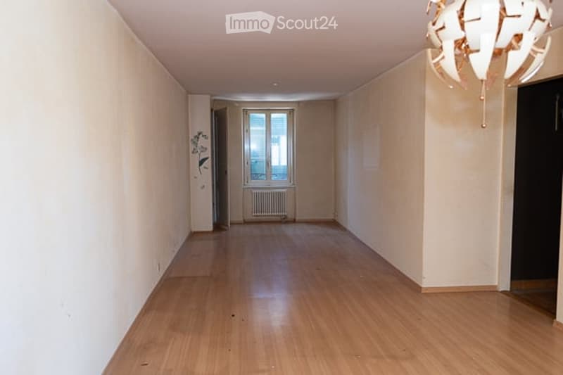Empty hallway with wooden flooring, white walls, and a chandelier hanging from the ceiling. There is a window at the end of the hallway with a radiator below it.
