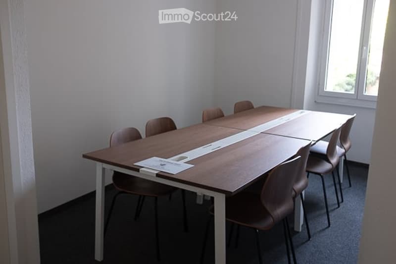 A small, modern meeting or conference room with a long wooden table and several chairs arranged around it. The room has white walls, a large window, and a carpeted floor.