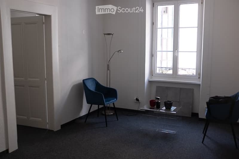 A small, modern living room with a blue armchair, a glass coffee table, and several cups on the table. The room has large windows and a floor lamp.