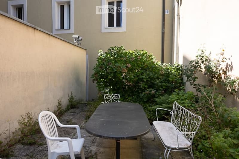 Small outdoor patio enclosed by a building wall and greenery, featuring a black rectangular table, a white plastic chair, and a decorative metal bench.