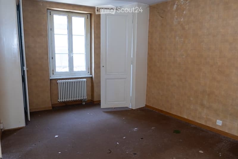 Empty room with bare walls, wooden floor, single window, white door, and radiator.
