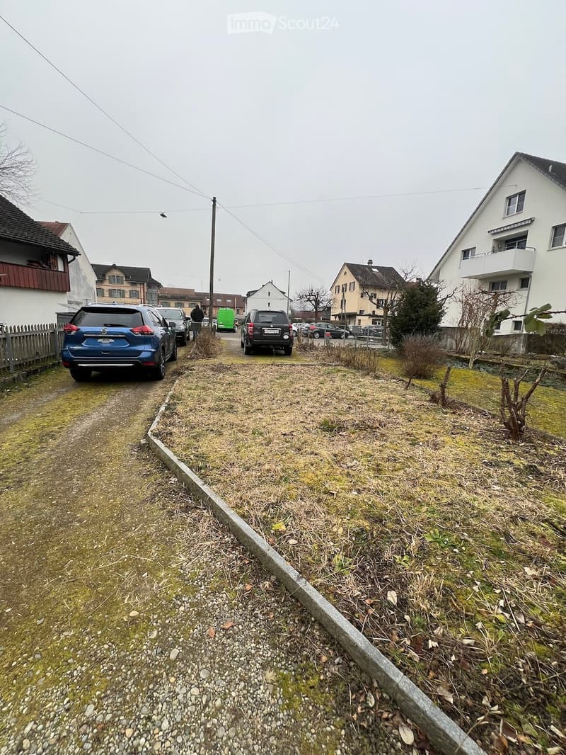 Residential street with parked cars, houses, and a grassy area with sparse vegetation.