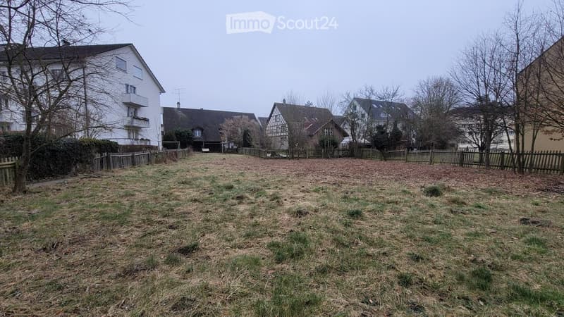 Empty garden area enclosed by a wooden fence, with bare ground and sparse grass, surrounded by residential buildings and trees.