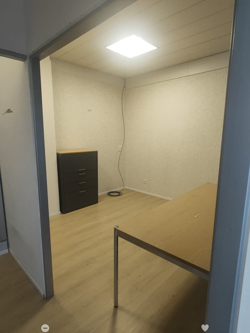 A small, unfurnished room with wooden flooring, a large wooden table, and a black cabinet with drawers.