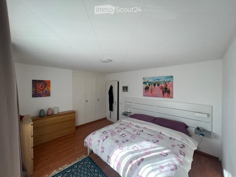 A well-lit bedroom with a double bed, nightstand, drawer, and decorative vases. The room features wooden flooring and white walls with artwork.