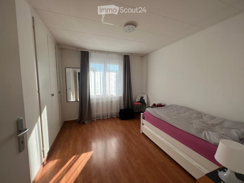 A small, well-lit bedroom with wooden flooring, a single bed with a pink blanket, a mirror, a small table, and a chair near the window with curtains.