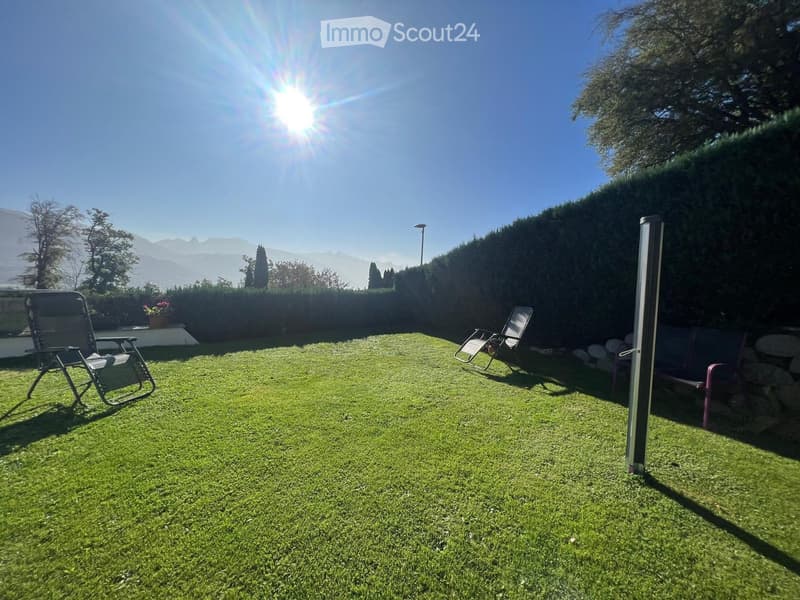 Well-maintained lawn with two chairs and a small table, surrounded by hedges and trees, with a scenic mountain view in the background.