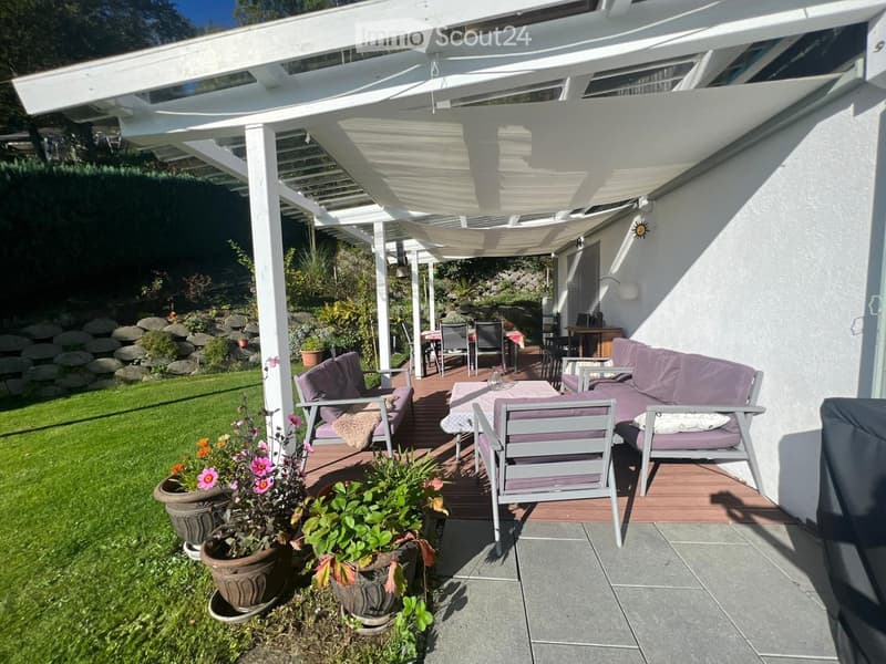 Outdoor patio with a covered pergola, sofa, chairs, and a dining table, surrounded by a well-maintained garden with potted plants and a stone wall.