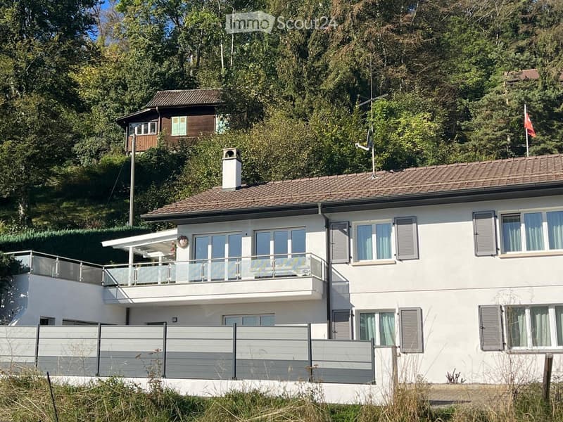 Two-story house with white exterior, tiled roof, multiple windows, and a balcony with glass railing. A smaller wooden extension is visible on the left side.