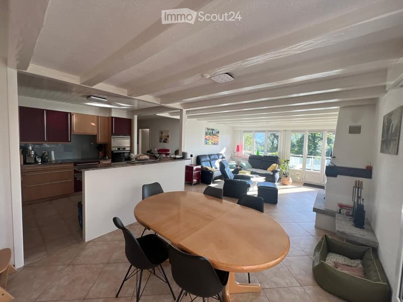 Open-plan living and dining area with a round wooden dining table, black chairs, a sofa, a fireplace, and a kitchen area with cabinets and appliances.