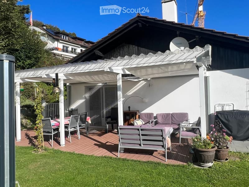 A well-maintained garden with a covered patio area, featuring a sofa, chairs, a bench, a dining table, and flower pots. The patio is connected to a house with a white exterior and a satellite dish on the roof.