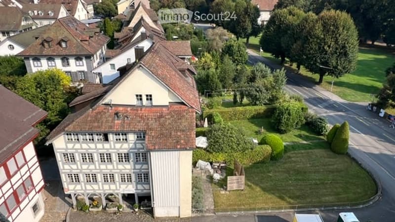 Aerial view of a traditional Swiss building with a red-tiled roof and white walls, surrounded by a well-maintained garden and neighboring houses.