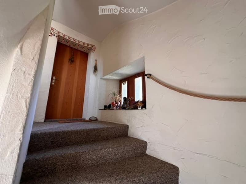 A narrow hallway with a carpeted staircase leading upwards, featuring a wooden door on one side and a small window with a decorative curtain on the other. The walls are textured white plaster.