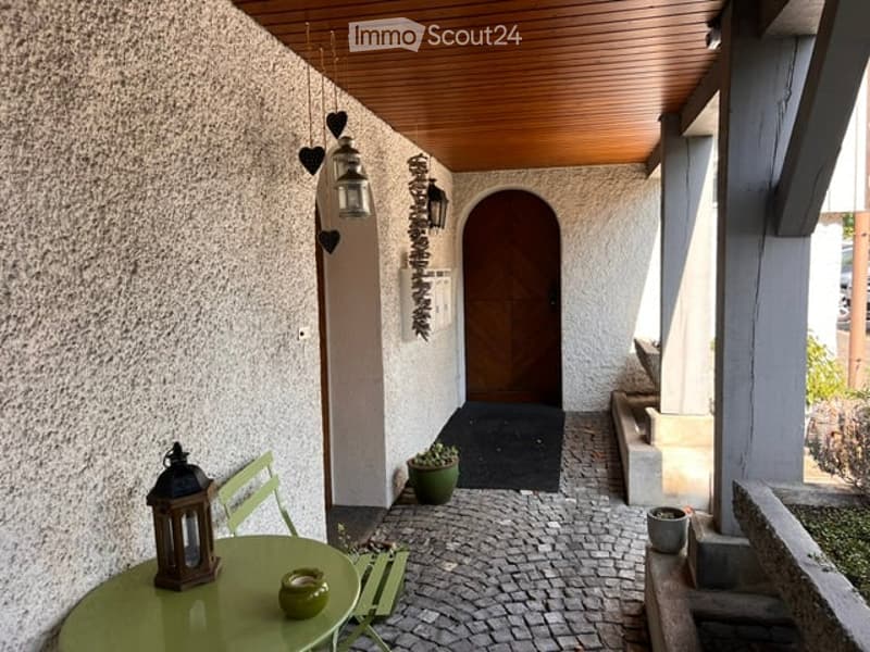 A small, cozy balcony with a wooden ceiling, green table and chairs, a lantern, and a potted plant near the entrance door.