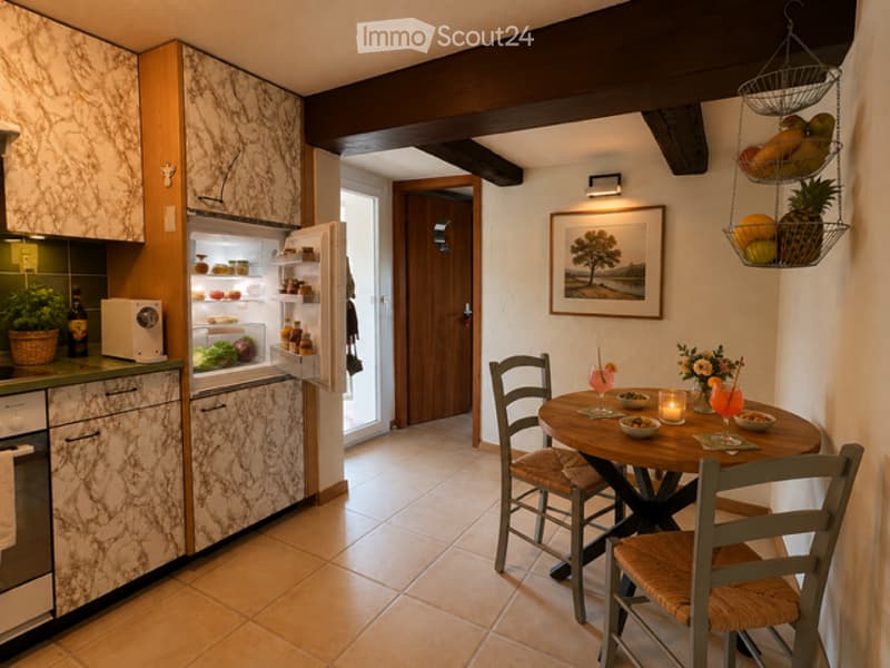 A cozy kitchen with marble countertops, a refrigerator, microwave, and a small dining area with a round table and chairs. A fruit basket and a flower vase are on the table, and a picture frame hangs on the wall.