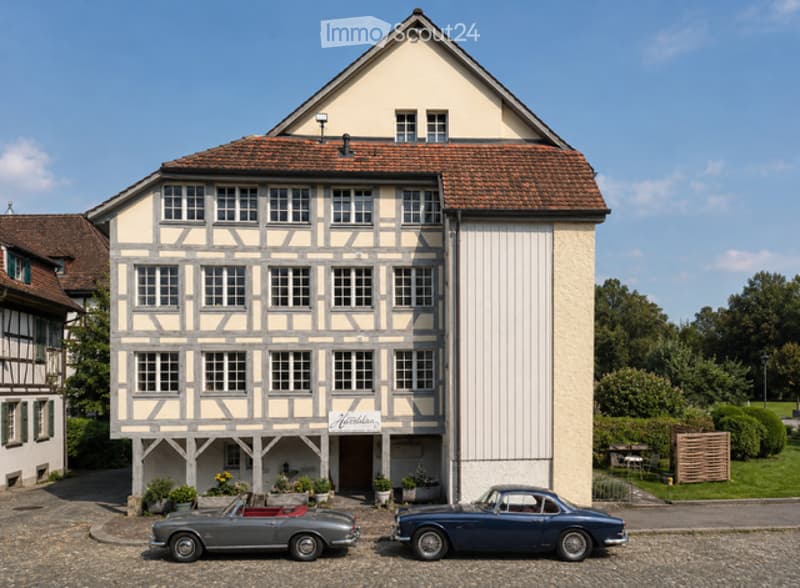 A two-story building with a classic facade, featuring a gabled roof and multiple windows. Two vintage cars are parked in front of the building.