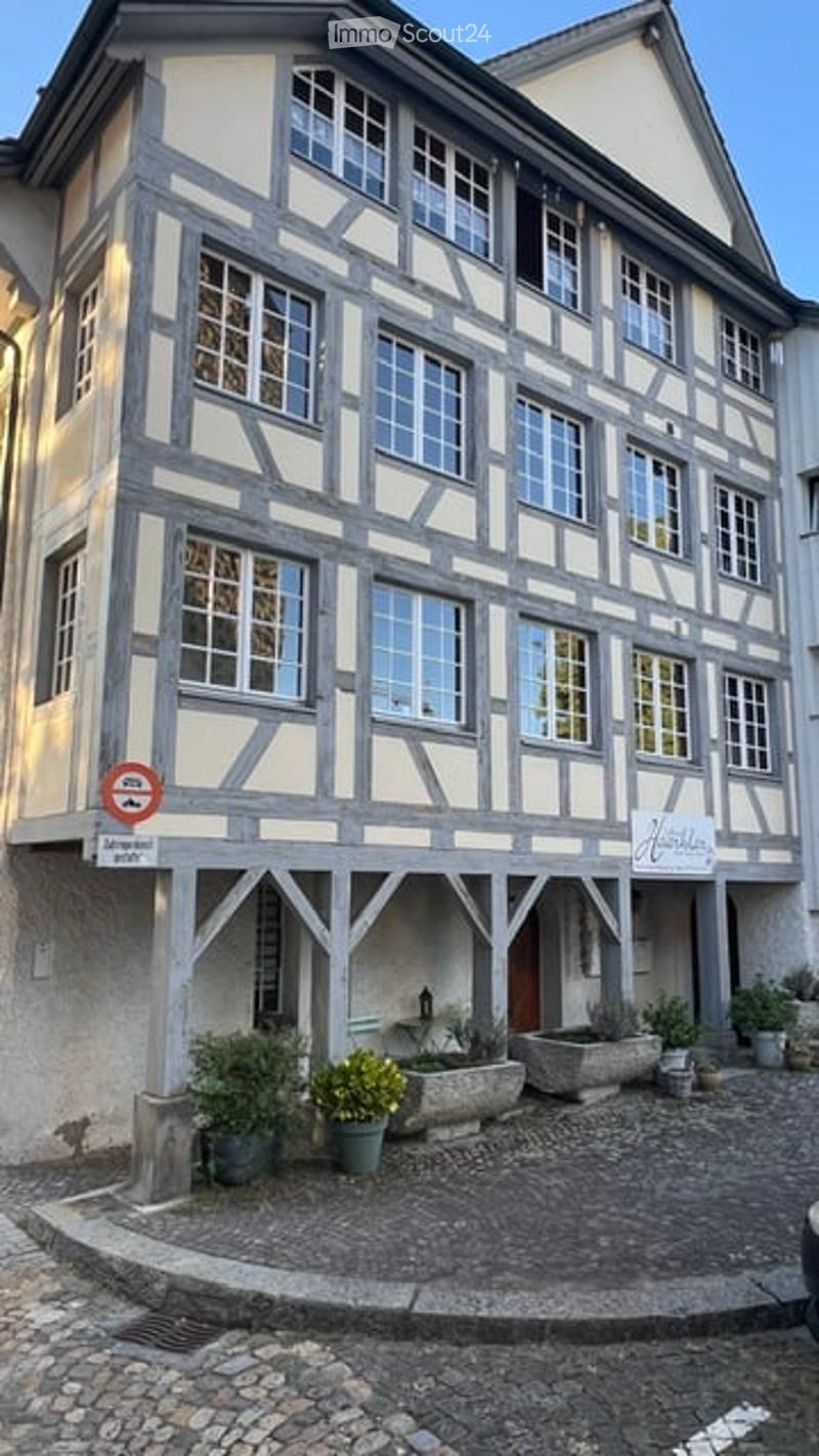 A traditional half-timbered building with white plaster and dark wooden beams, featuring multiple windows and a small garden area with potted plants and a stone planter in front.