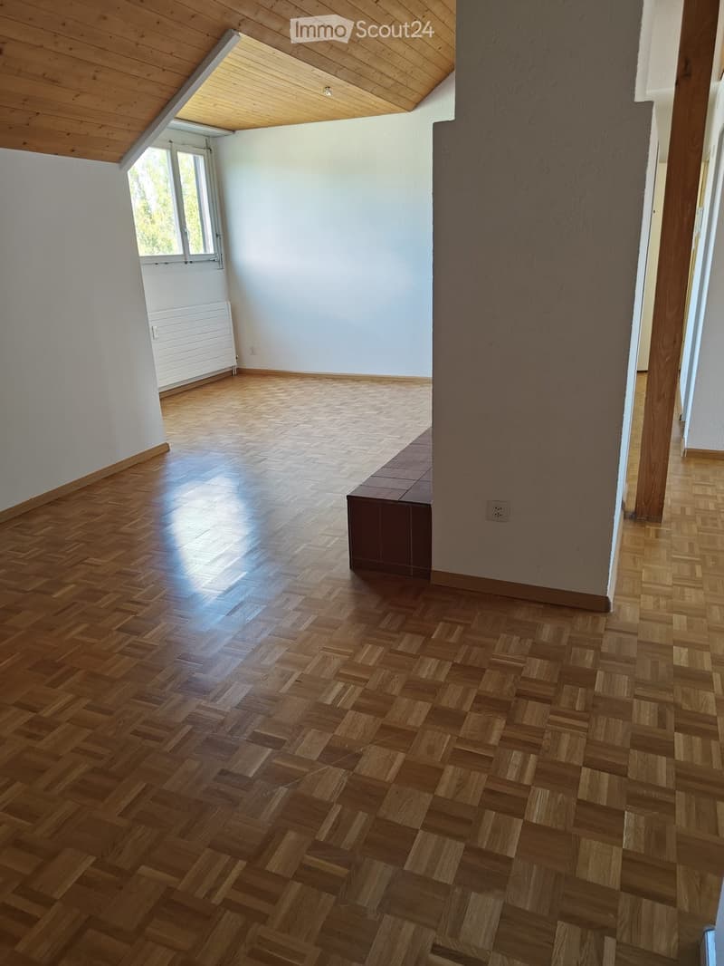Empty room with wooden herringbone flooring, white walls, and a slanted wooden ceiling. Features a window with white frame and radiator below, and a small dark-colored bench against one wall.