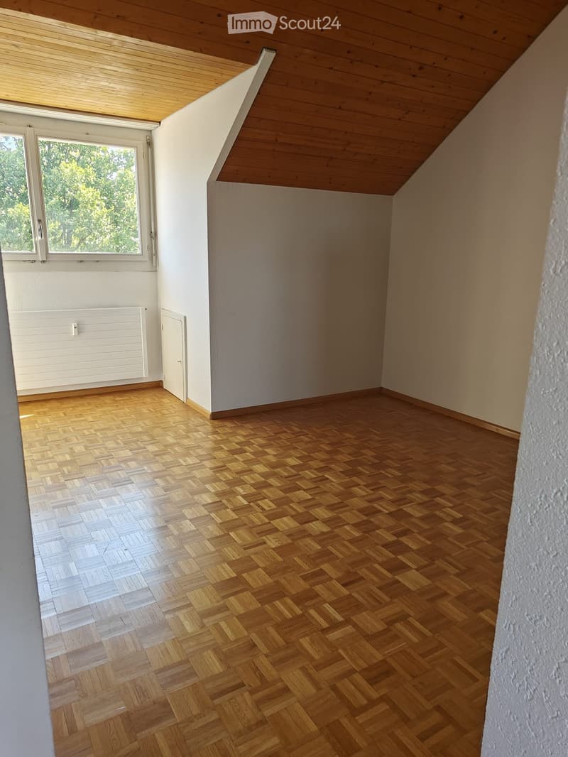 Empty room with wooden herringbone flooring, white walls, and a slanted wooden ceiling. Features a window with white frame and a radiator below it.
